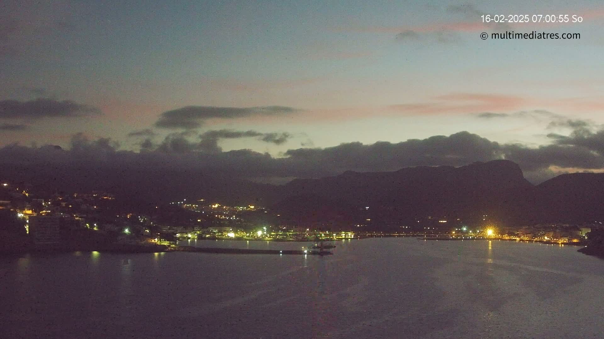 A nighttime coastal scene shows a town with lights nestled against dark mountains under a dusky sky with clouds.