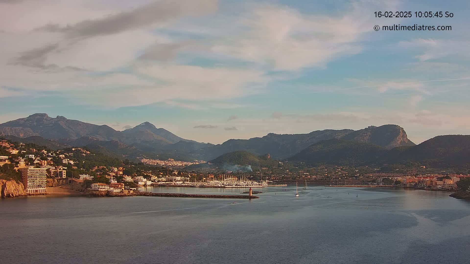 A coastal town nestled between mountains under a partly cloudy sky, with a marina filled with boats in the foreground.