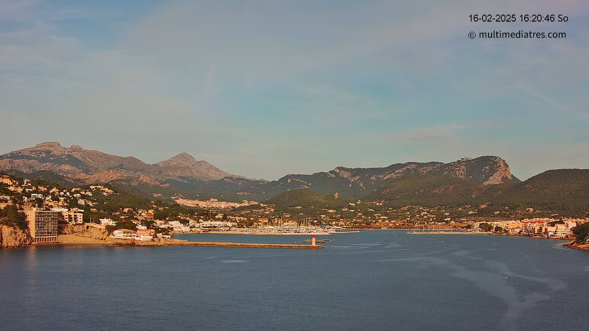 A coastal town nestled at the base of mountains, with a calm body of water in the foreground, under a clear, light-blue sky.