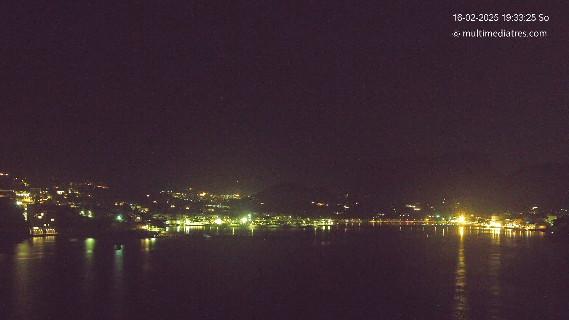 A nighttime view of a city situated on a hillside next to a calm body of water, reflecting the city lights.