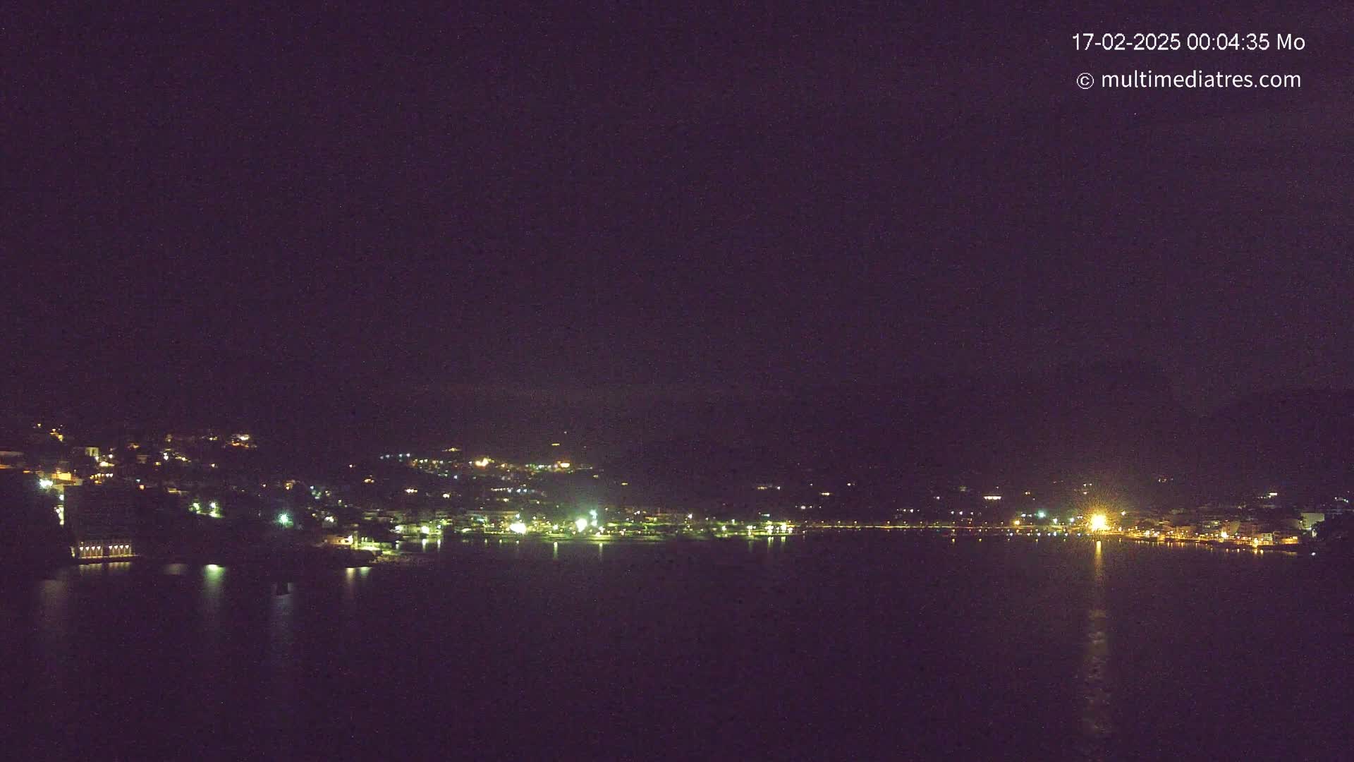 A nighttime view shows a town's lights scattered across a hillside beside a dark body of water.