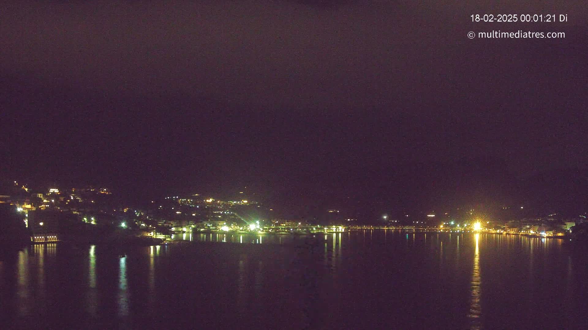 A nighttime view of a city's lights reflected on a calm body of water under a dark sky.