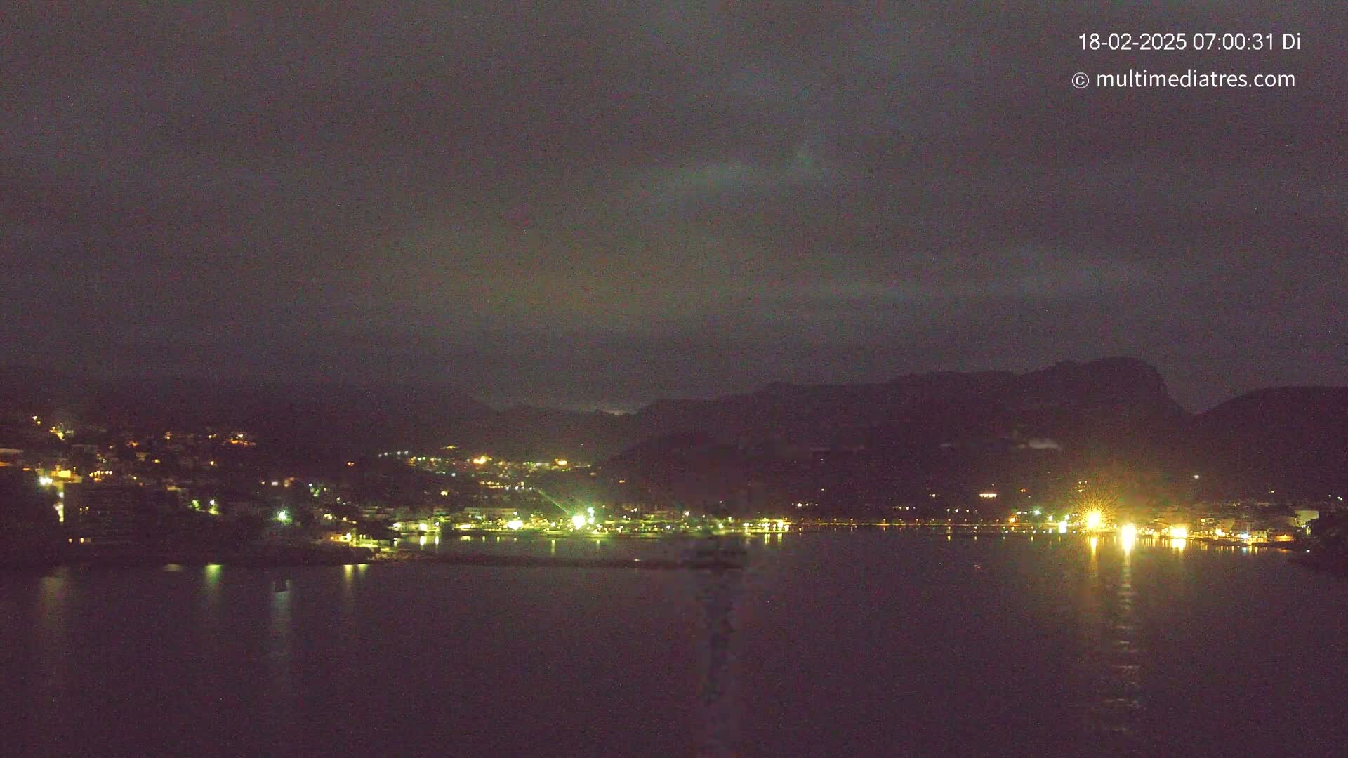 A nighttime view of a city situated along a calm body of water, nestled between dark hills under a mostly clear, dark sky.