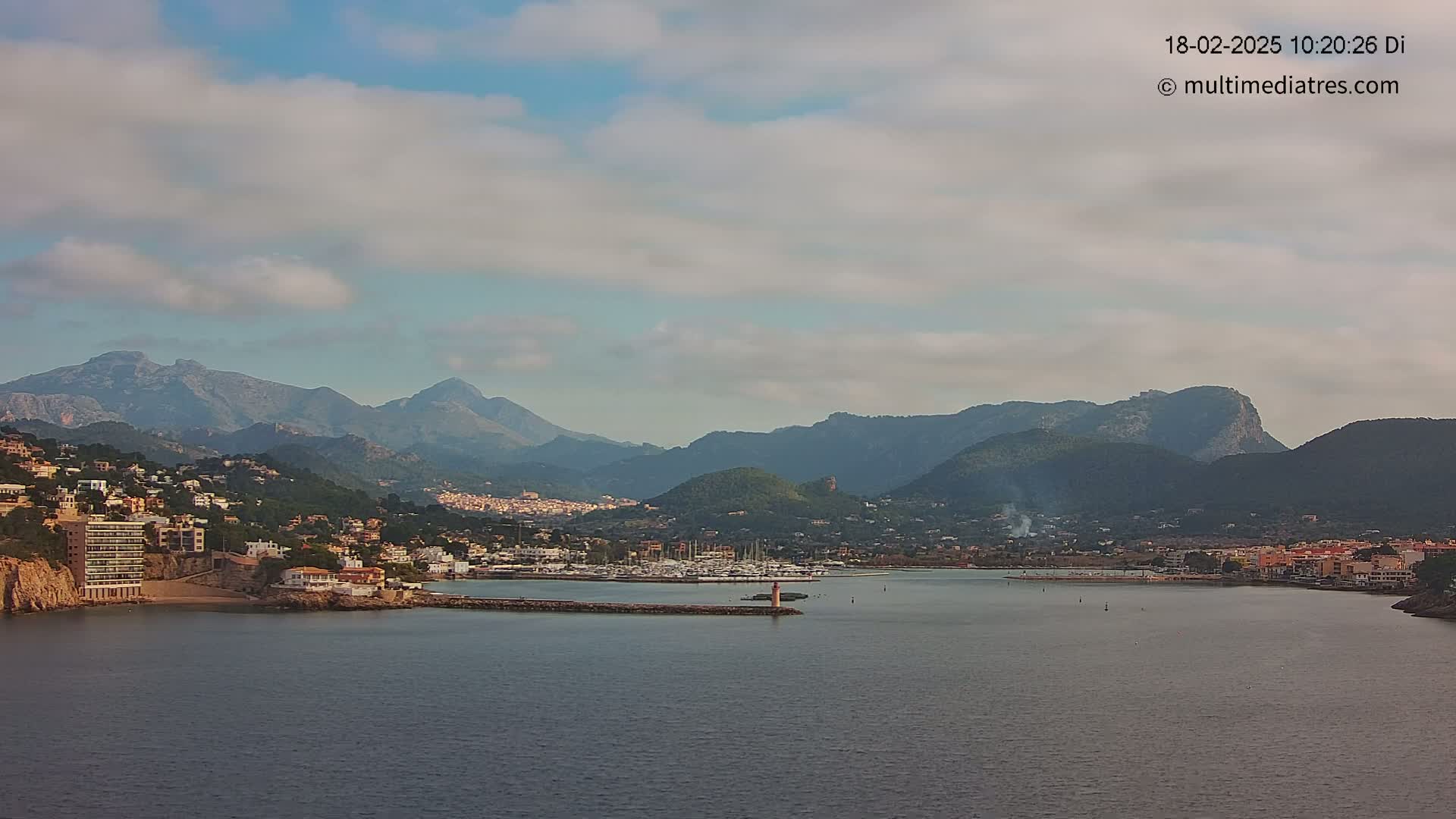 A coastal town nestled against a mountain range under a partly cloudy sky, with a calm bay filled with boats in the foreground.