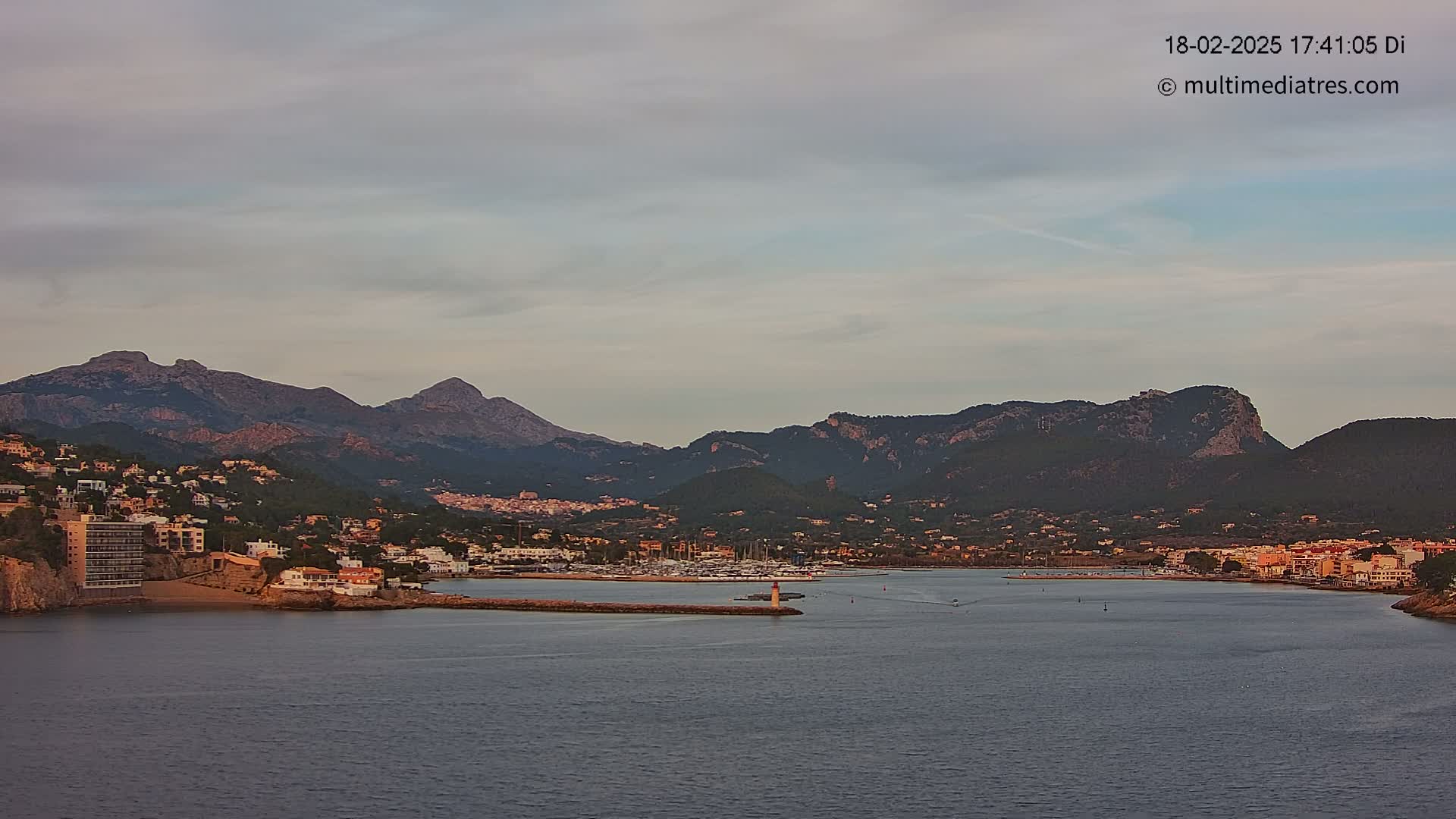 A coastal town nestled at the base of a mountain range is visible under a mostly cloudy sky, with a calm body of water separating the viewer from the town and mountains.