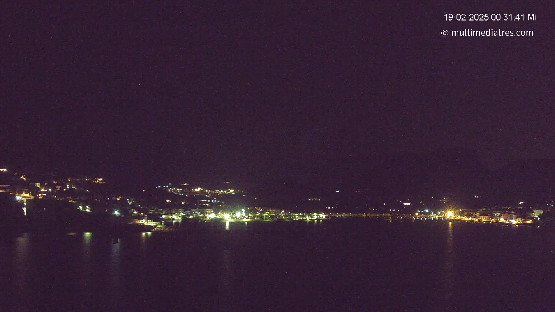 A nighttime view of a coastal town with its lights reflected on a calm body of water under a dark sky.