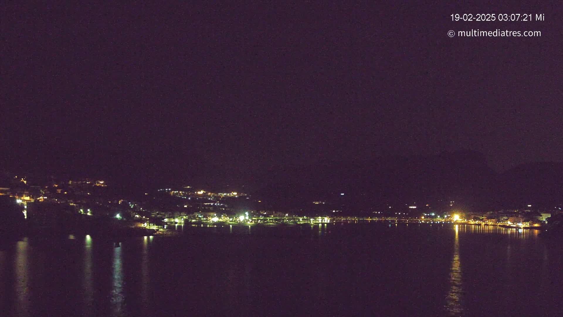 A nighttime view shows a calm body of water reflecting the lights of a town nestled on a hillside under a dark sky.