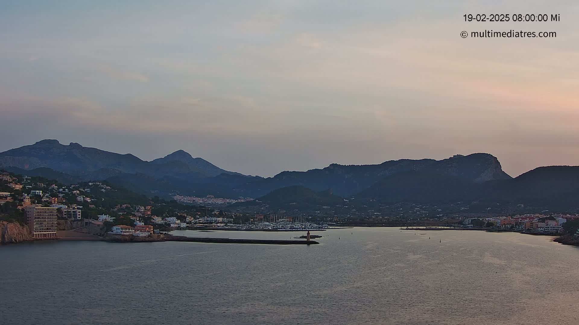 A coastal town nestled at the base of dark mountains is seen under a pale, dusky sky, with a calm body of water separating the town from a harbor filled with boats.