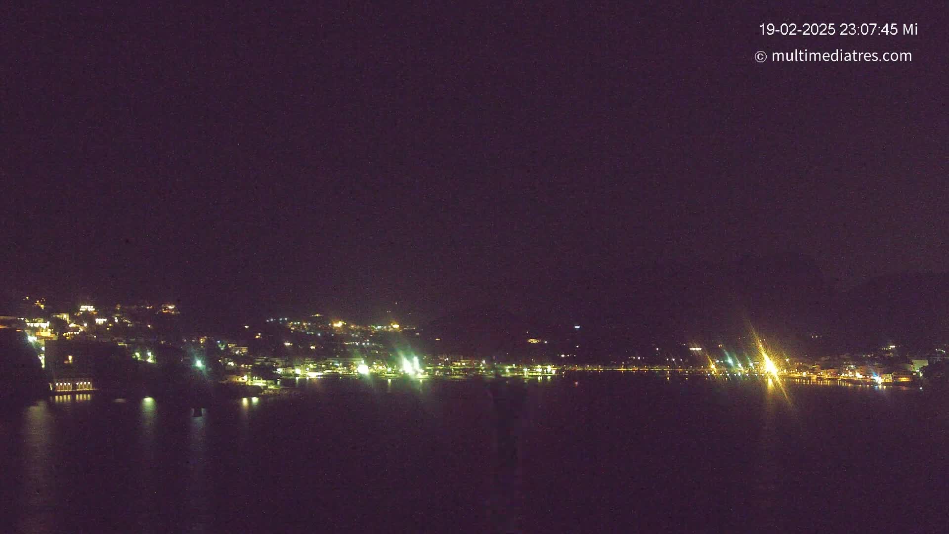 A nighttime view of a town's lights reflected on a calm body of water under a clear, dark sky.