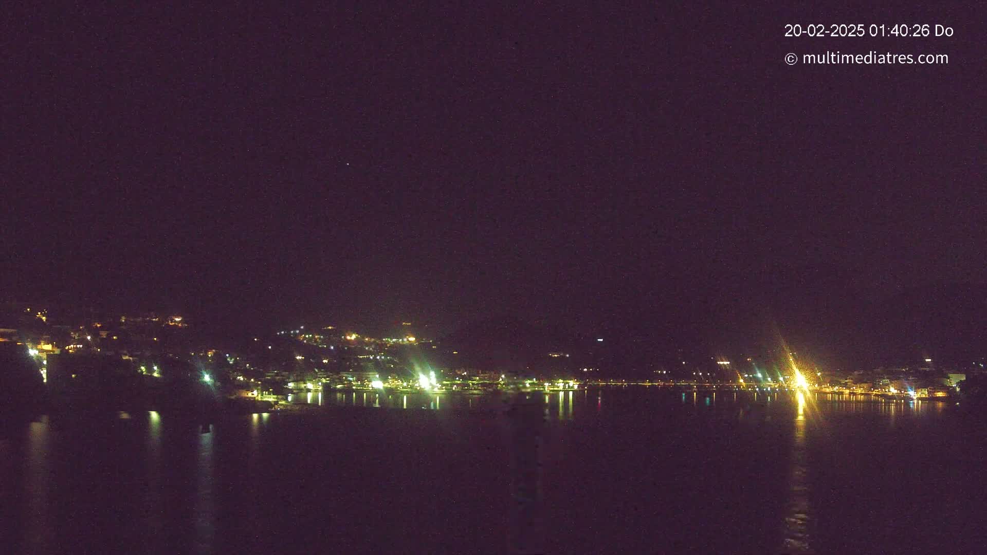 A nighttime view of a coastal town's lights across a calm body of water under a dark, clear sky.