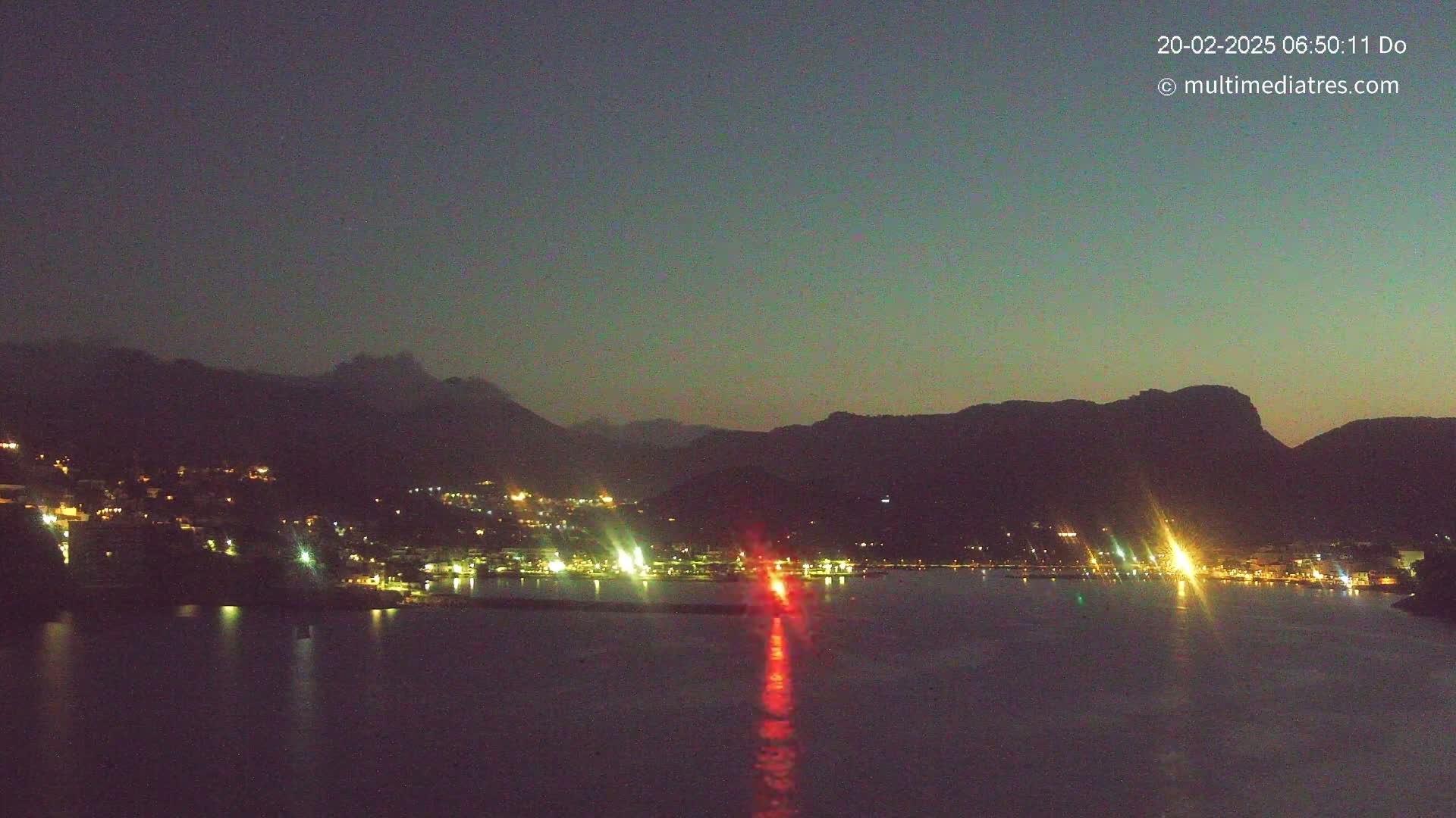 A nighttime view of a coastal town nestled between dark mountains, illuminated by scattered lights, with a calm body of water reflecting the lights and a single bright red light in the center.