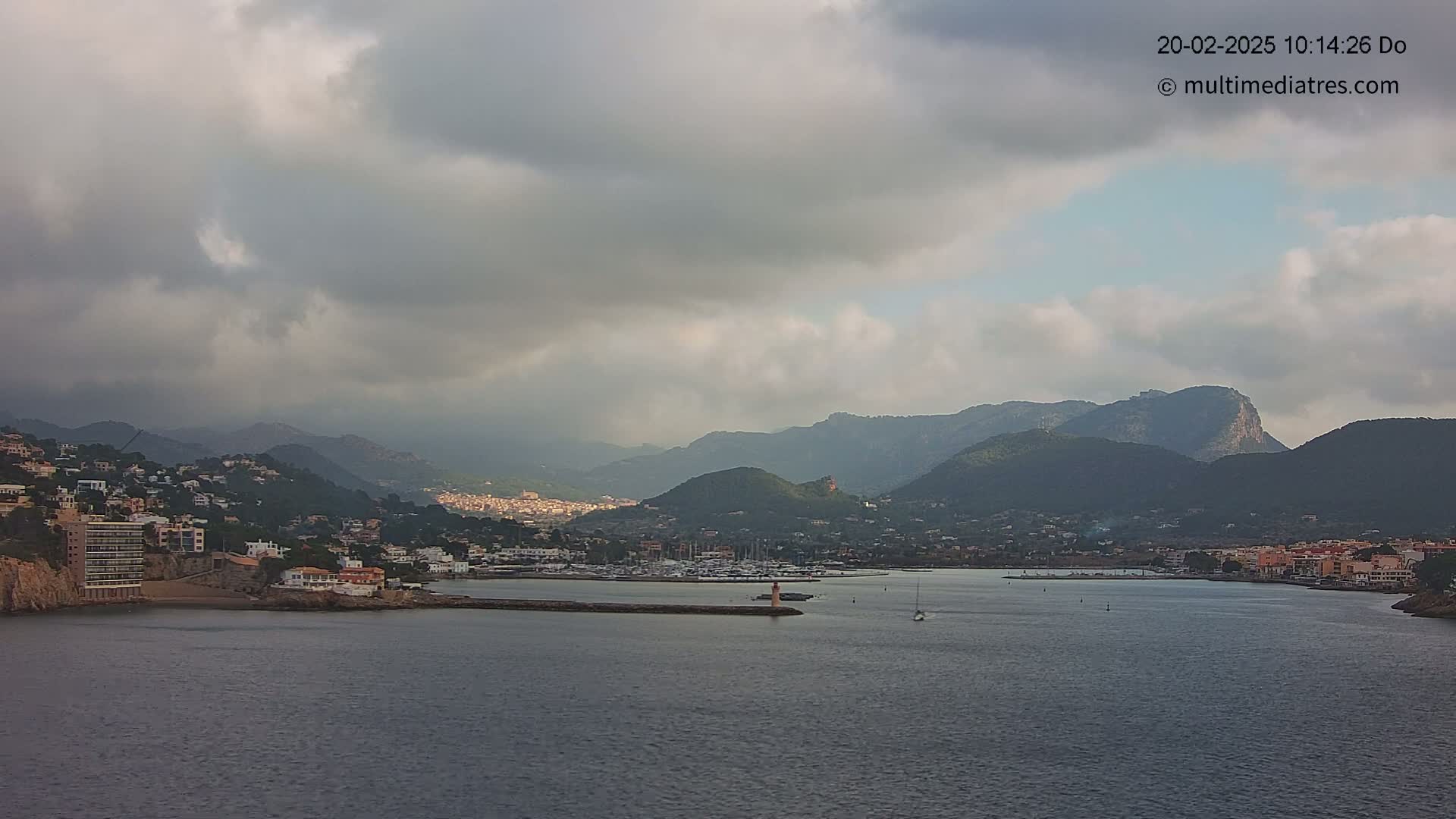 A coastal town nestled between mountains is partially obscured by an overcast sky, with a calm body of water in the foreground containing a marina.