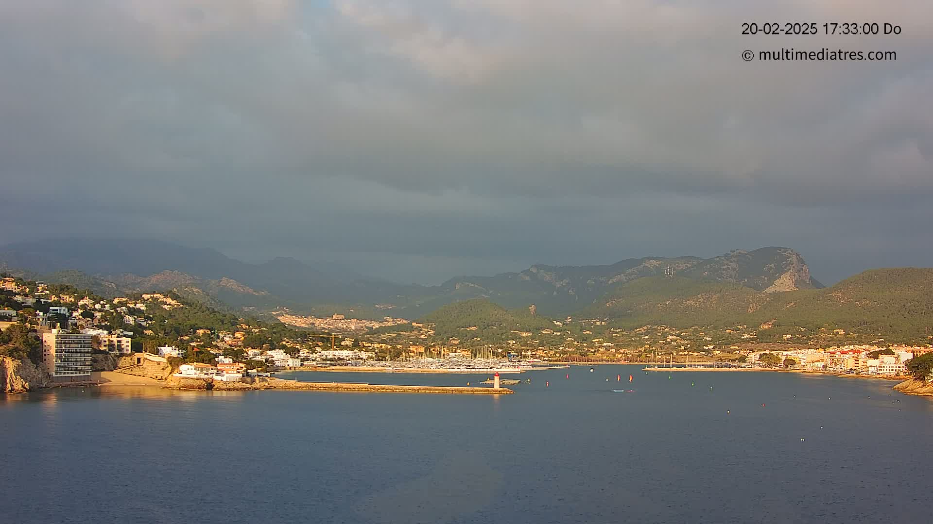 A coastal town nestled between mountains under a cloudy sky, with a marina filled with boats.