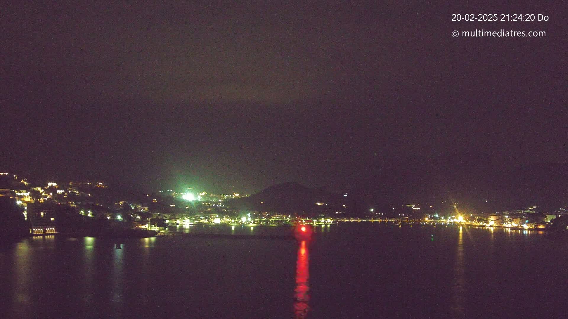 A nighttime view of a town situated on a hill overlooking a dark body of water, with a single red light visible on the water's surface.