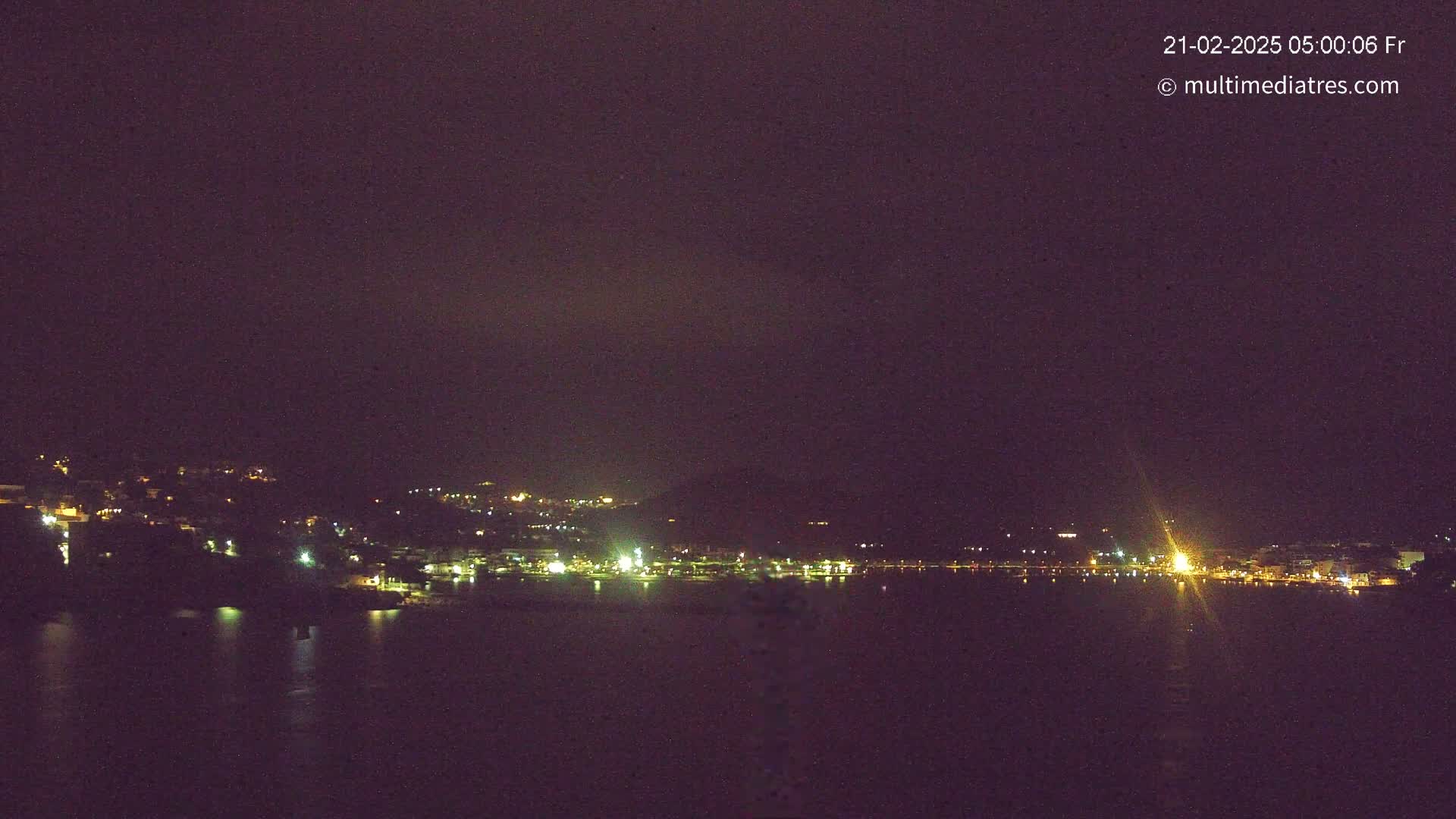 A nighttime view of a town's lights across a dark body of water under a mostly clear, dark sky.