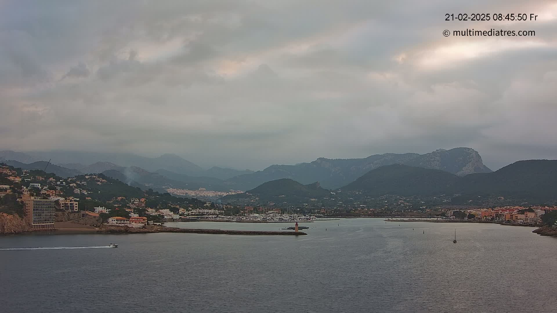 A coastal town nestled between mountains is seen under an overcast sky, with a calm body of water containing a few boats in the foreground.