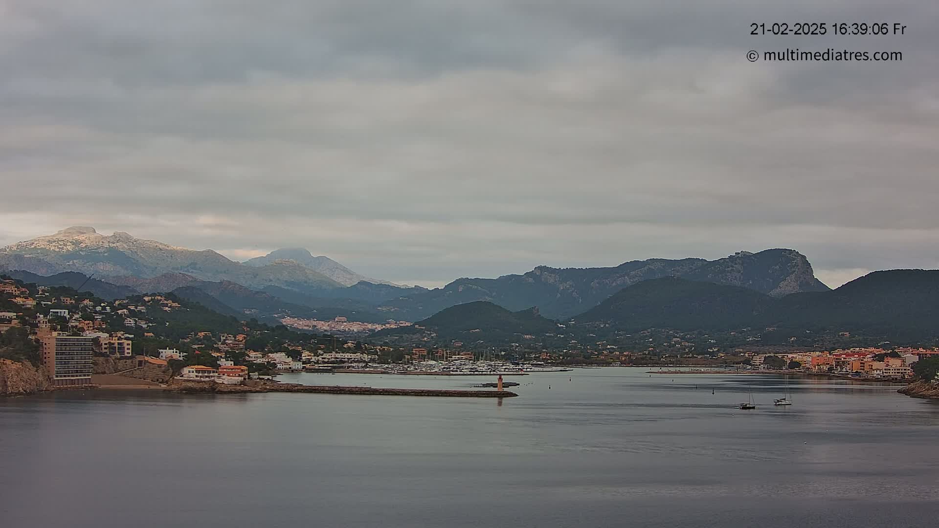 A coastal town nestled between mountains under an overcast sky, with a calm harbor containing several boats.