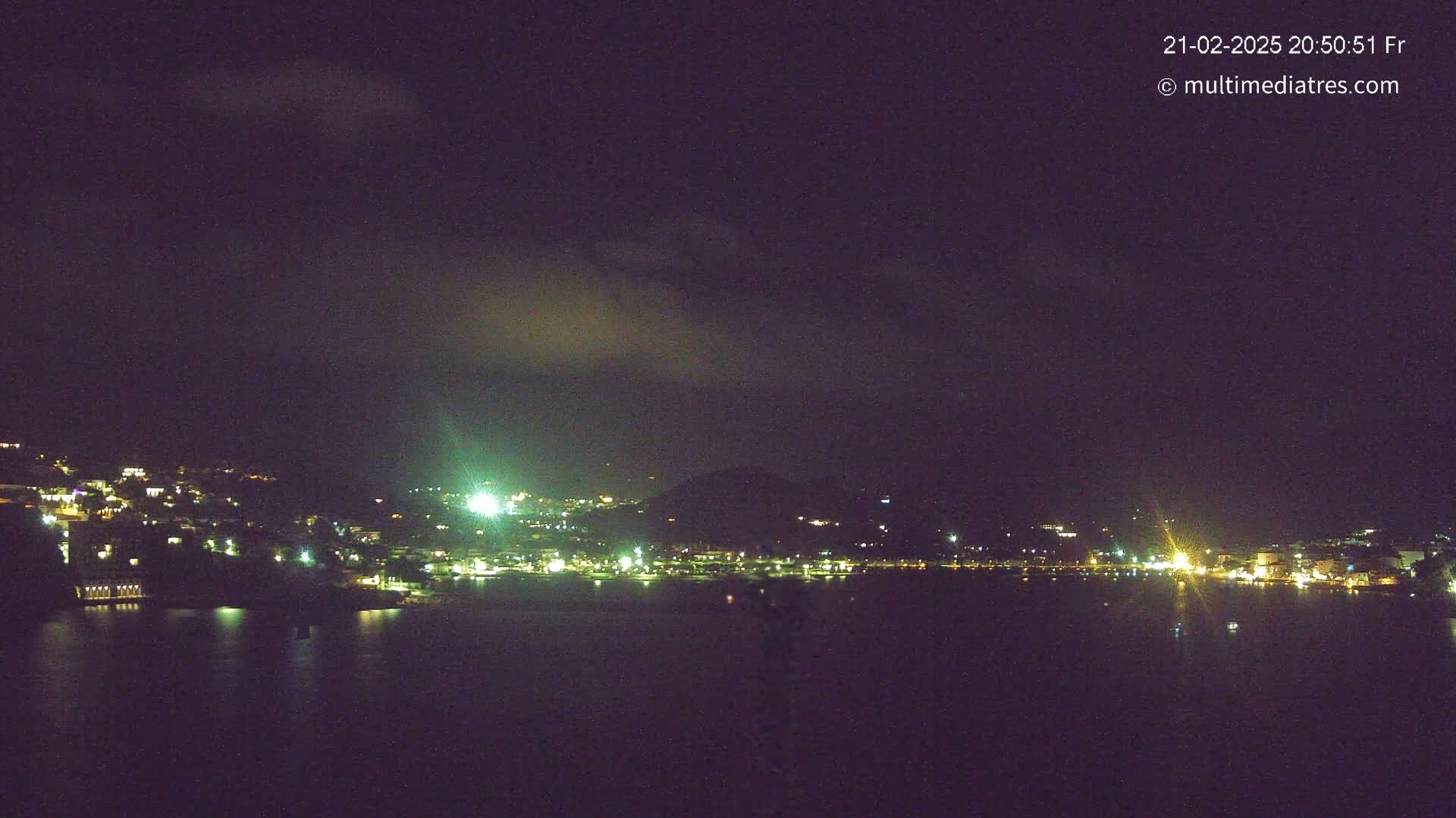 A nighttime view of a coastal town with its lights reflecting on a calm body of water under a mostly cloudy sky.