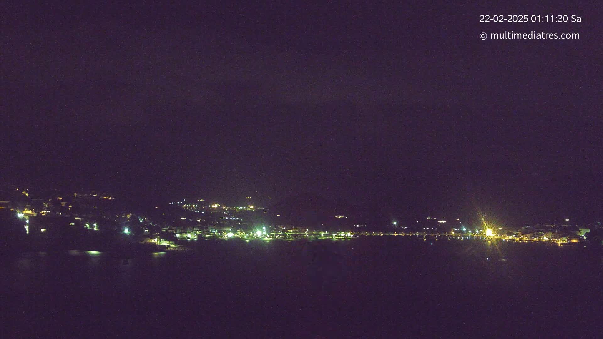 A nighttime view of a coastal town with scattered lights across a dark hillside and along a body of water under a clear, dark sky.