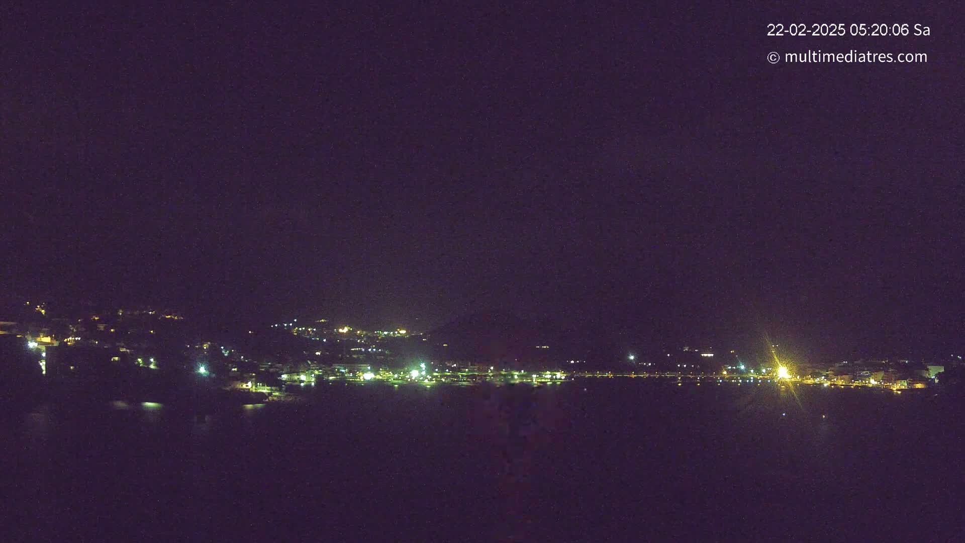 A nighttime long shot shows a coastal town with scattered lights along a dark body of water under a clear, dark sky.