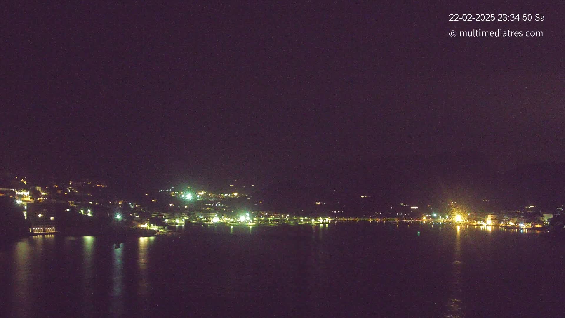 A nighttime view of a town's lights spread across a hillside and reflected in a calm body of water under a dark sky.