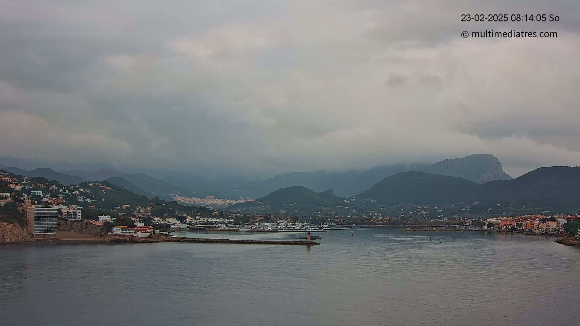 A coastal town nestled between dark mountains under a cloudy sky, with a calm body of water separating the town from a breakwater with boats moored beyond it.
