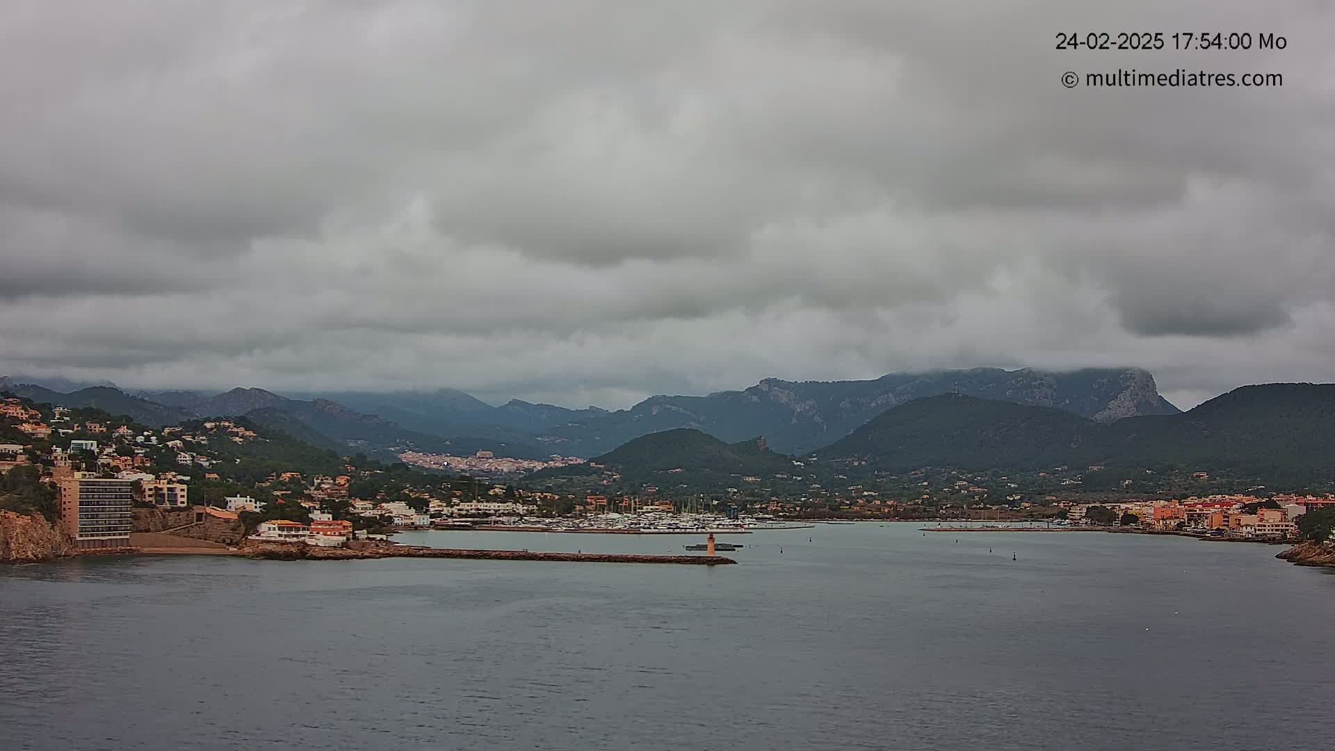 A coastal town nestled between mountains under a cloudy sky, with a calm harbor filled with boats.