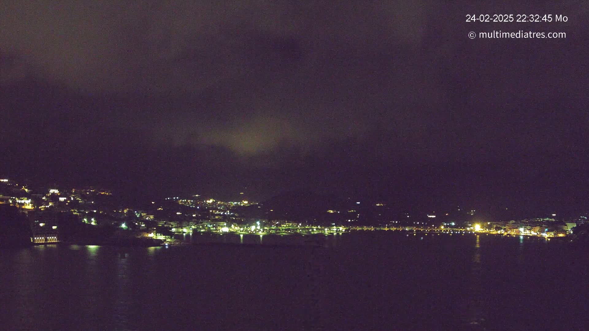 A nighttime view of a coastal town illuminated by lights, under a dark, cloudy sky.