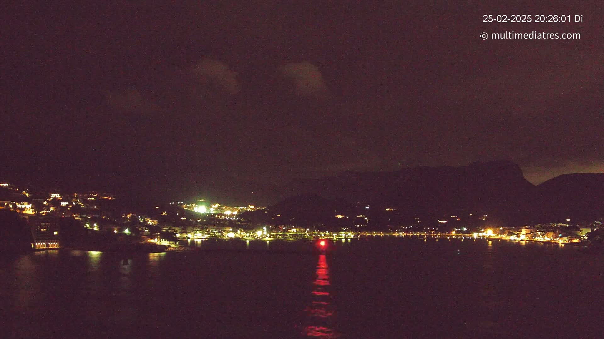 A nighttime view of a coastal town illuminated by lights, nestled against a dark mountain under a mostly clear, dark sky.