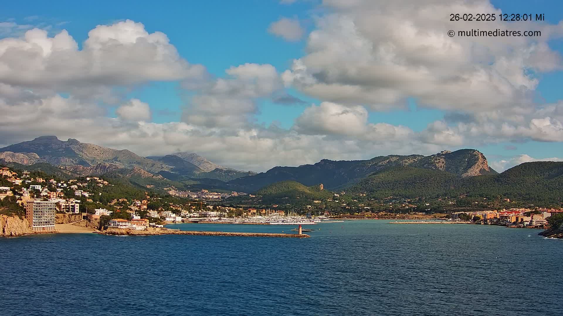 A partly cloudy day shows a coastal town nestled between mountains and a calm, dark blue sea with a marina filled with boats.