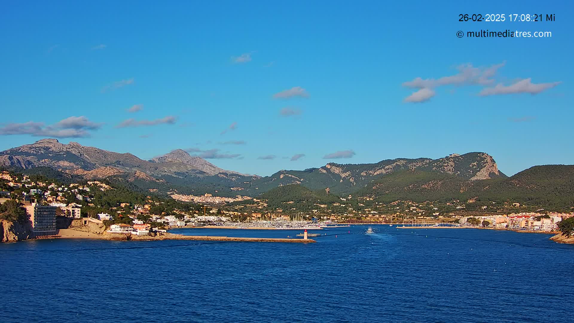 A coastal town nestled between mountains and a calm blue sea under a mostly clear blue sky with a few scattered clouds.