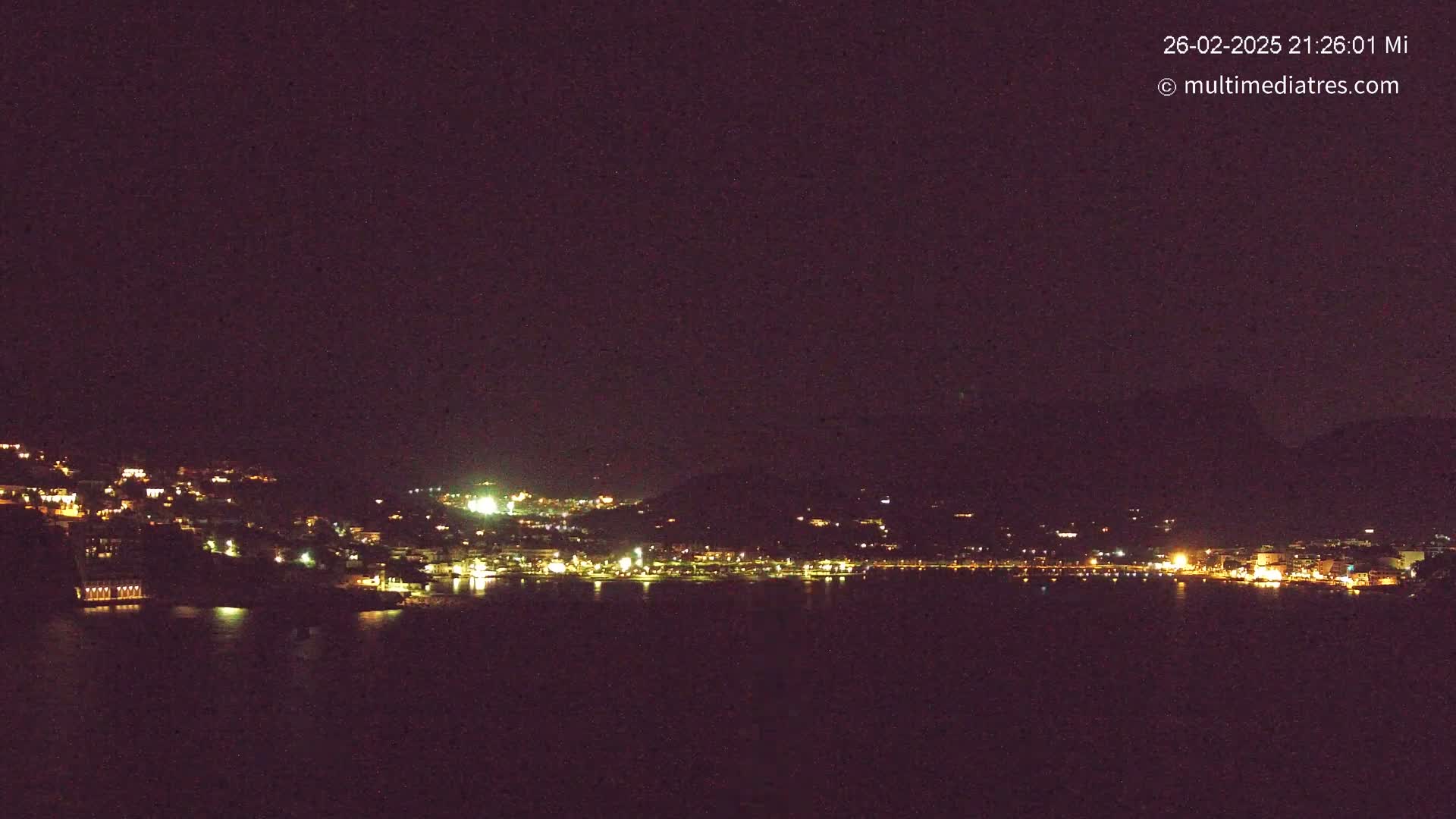A nighttime, long shot shows a town’s lights illuminated along a dark body of water and hillside, under a clear, dark sky.