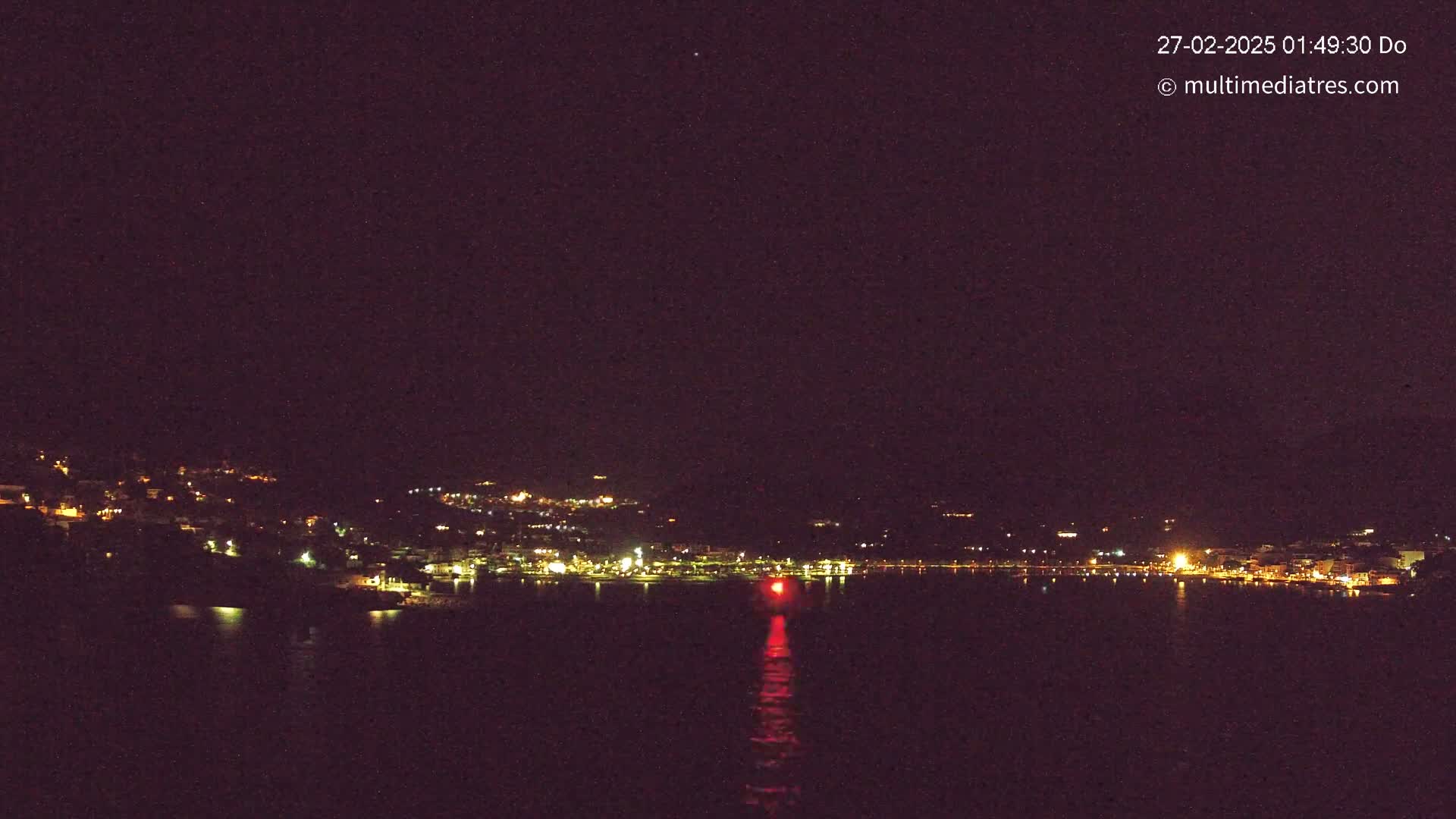 A nighttime view of a coastal town with its lights reflecting on calm dark water under a clear, starlit sky.