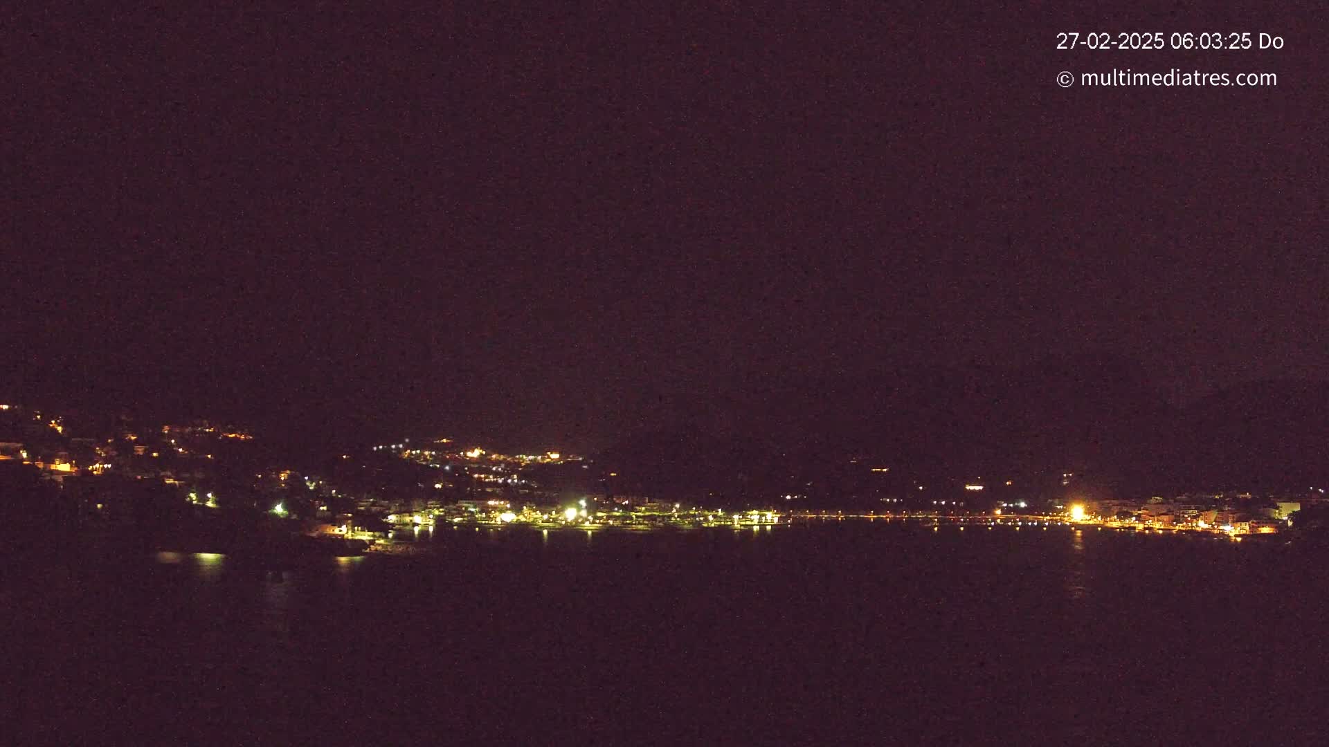 A nighttime view of a coastal town illuminated by scattered lights, across a dark body of water under a clear, dark sky.