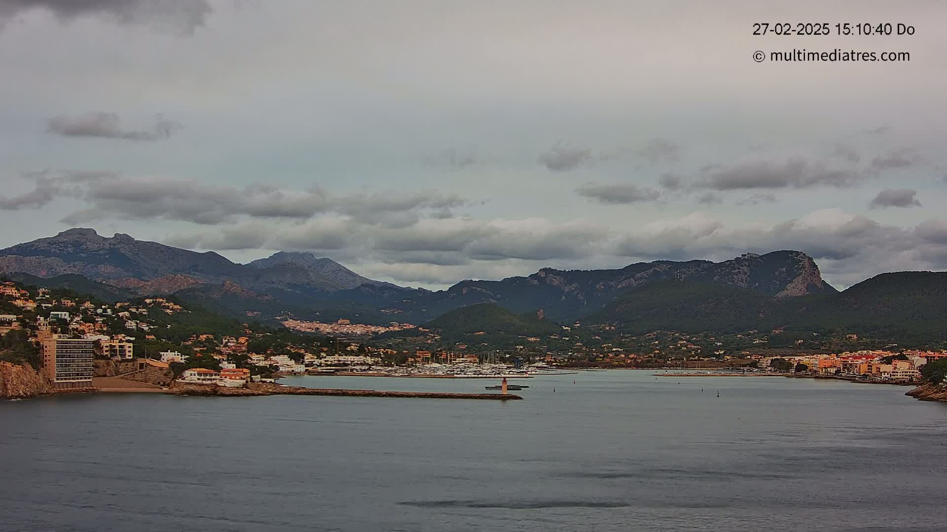 A coastal town nestled between a calm bay and a mountain range under a cloudy sky.