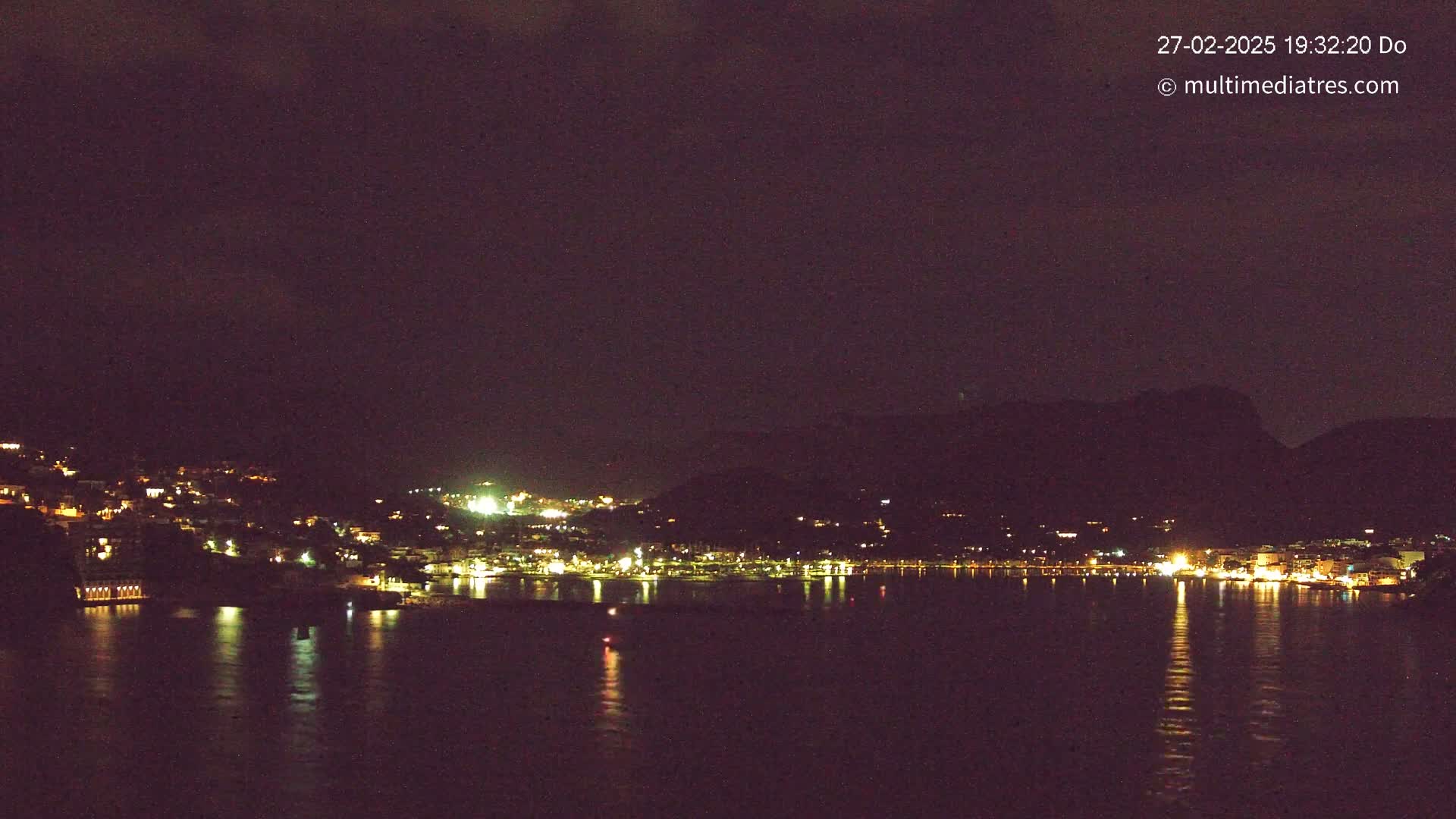 A nighttime view of a coastal town illuminated by lights, reflected in calm water under a dark sky.
