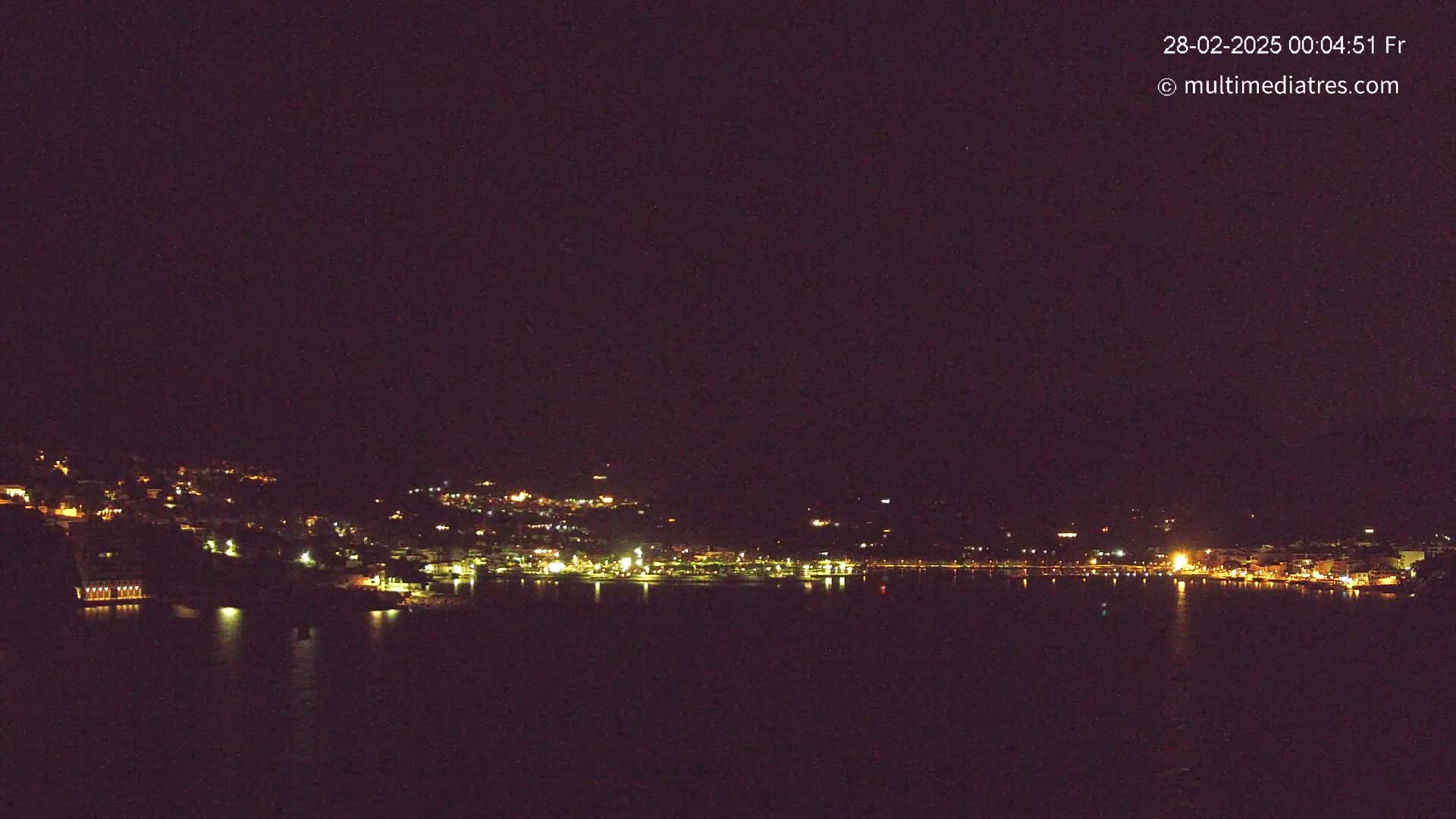 A nighttime view of a coastal town with its lights reflecting on a dark body of water under a clear, dark sky.