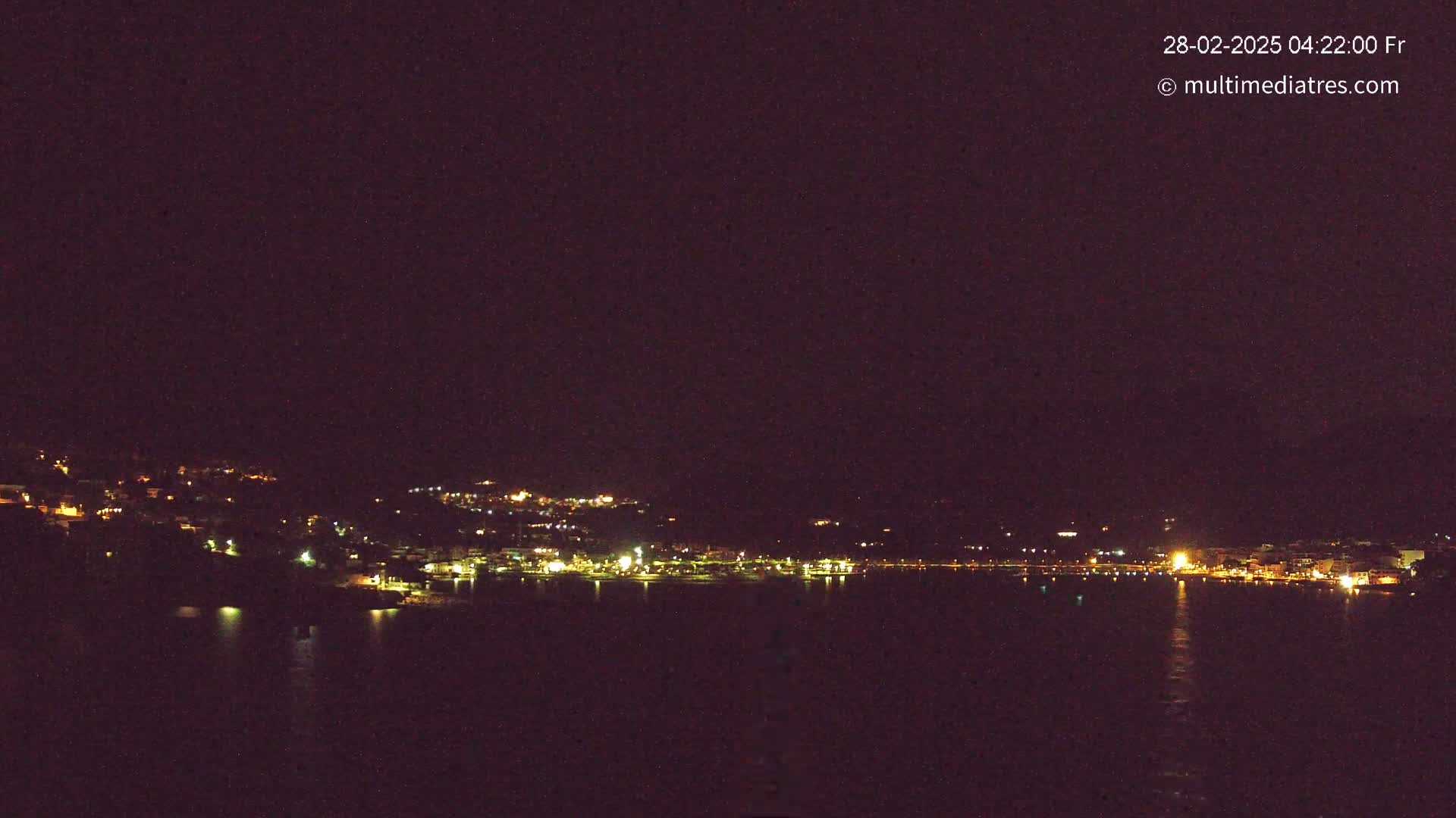 A nighttime, dark-sky view of a coastal town illuminated by lights, situated across a body of dark water.