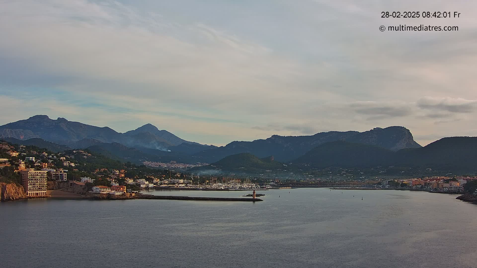 A coastal town nestled between mountains and a calm body of water under a mostly cloudy sky.