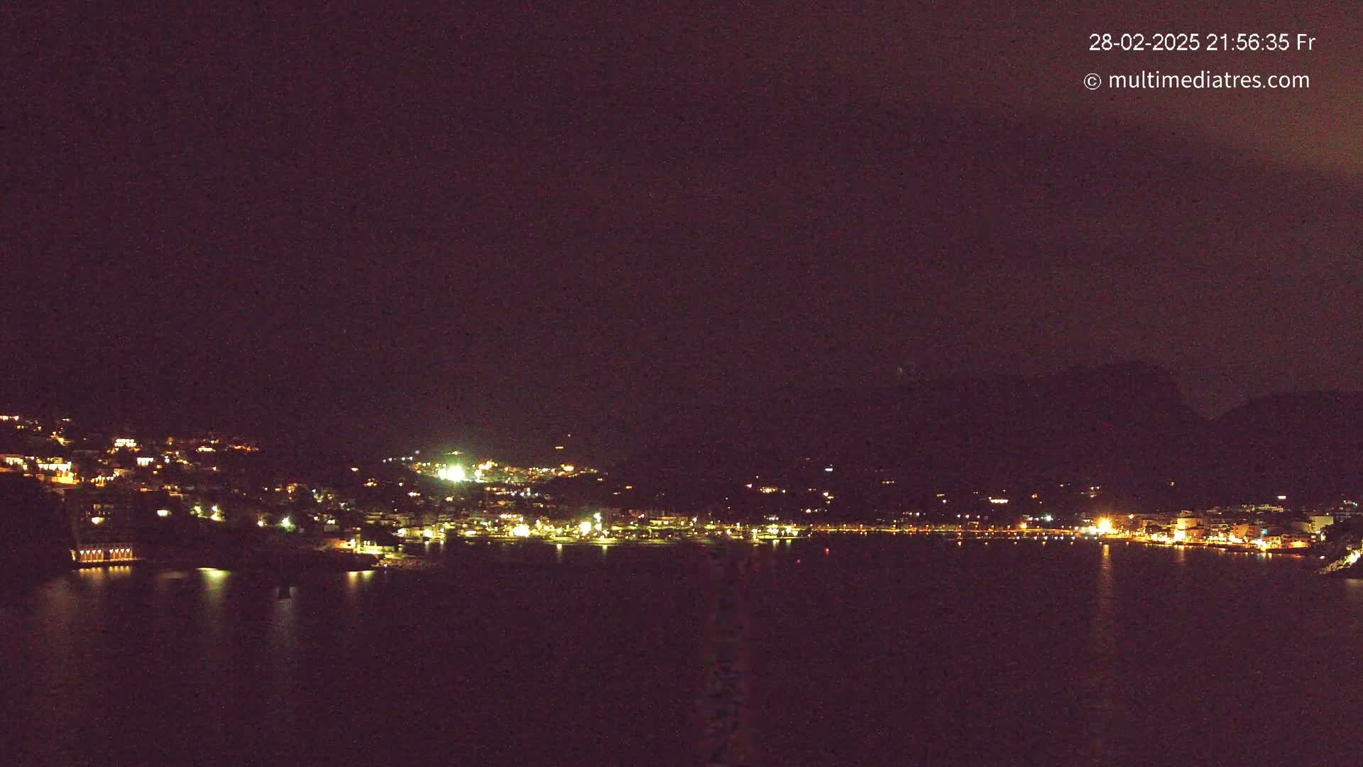 A nighttime view of a town situated on a body of water, nestled against a dark hillside under a clear, dark sky.