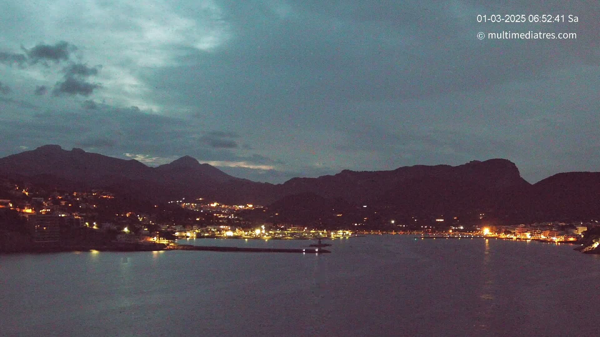 A nighttime view of a coastal town illuminated by lights, nestled between dark mountains under a dusky sky.