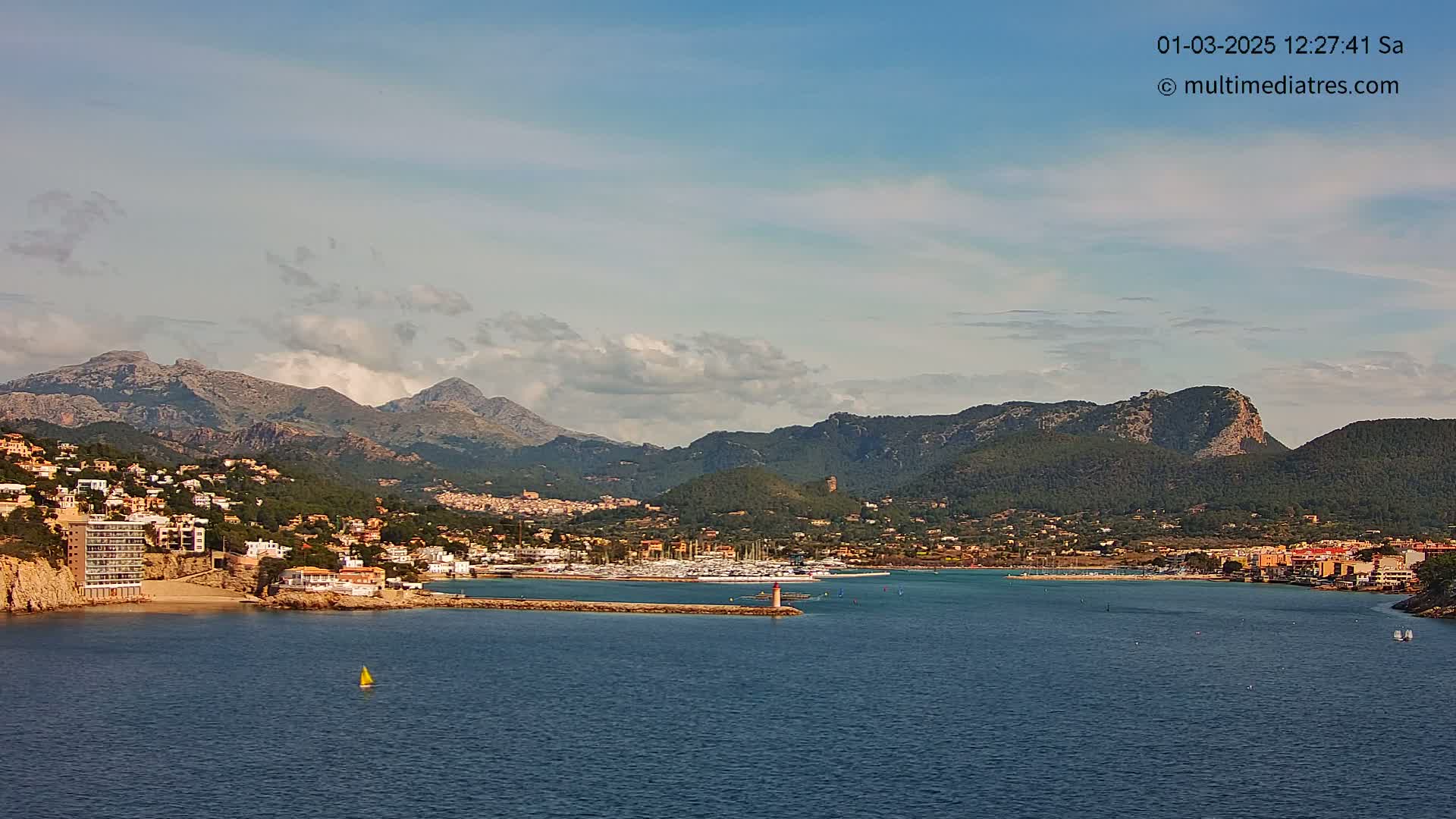 A coastal town nestled against a mountain range under a partly cloudy sky, with a marina filled with boats and a small sailboat on the calm water.