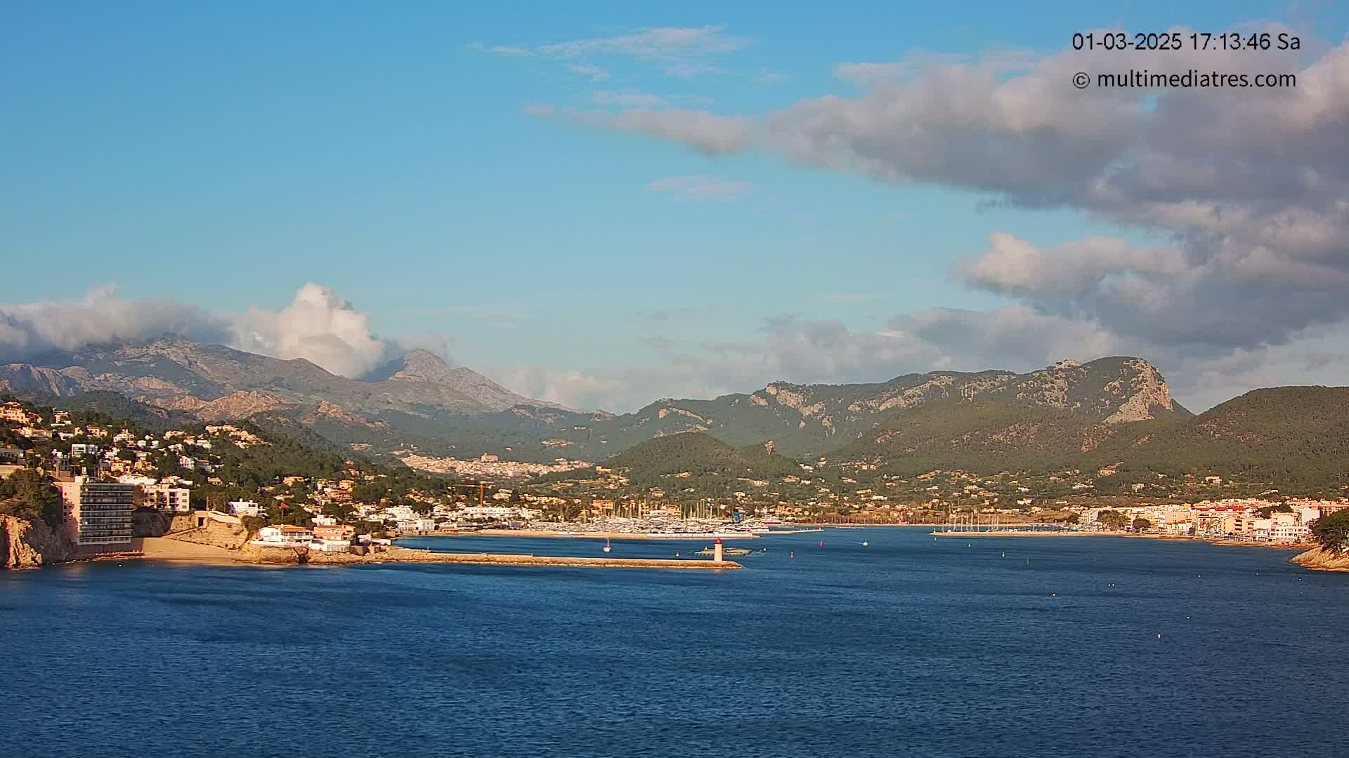 A coastal town nestled between mountains under a partly cloudy sky sits on a calm blue sea with a marina visible in the foreground.