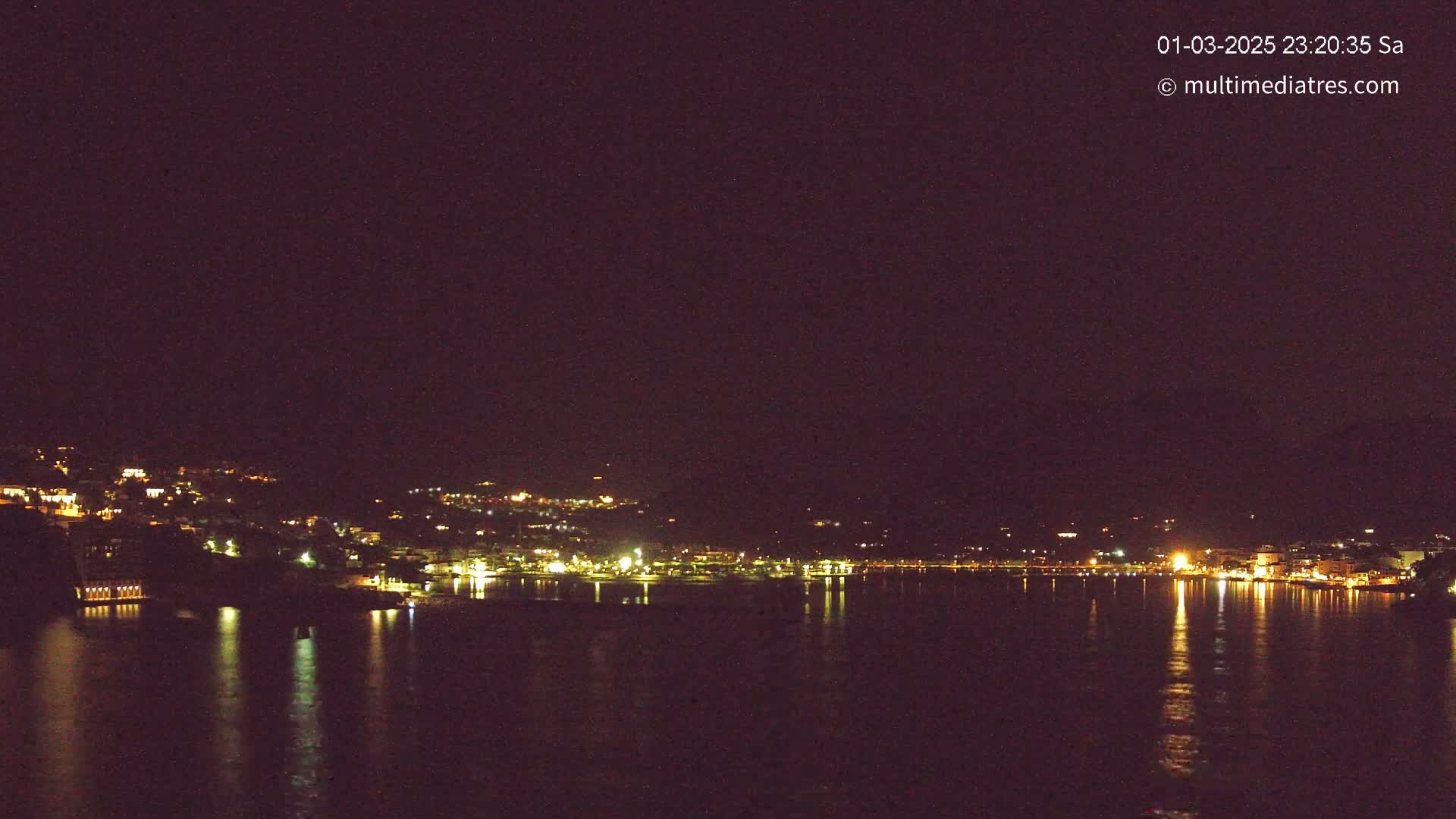 A nighttime view of a city's lights reflected on calm water under a clear, dark sky.
