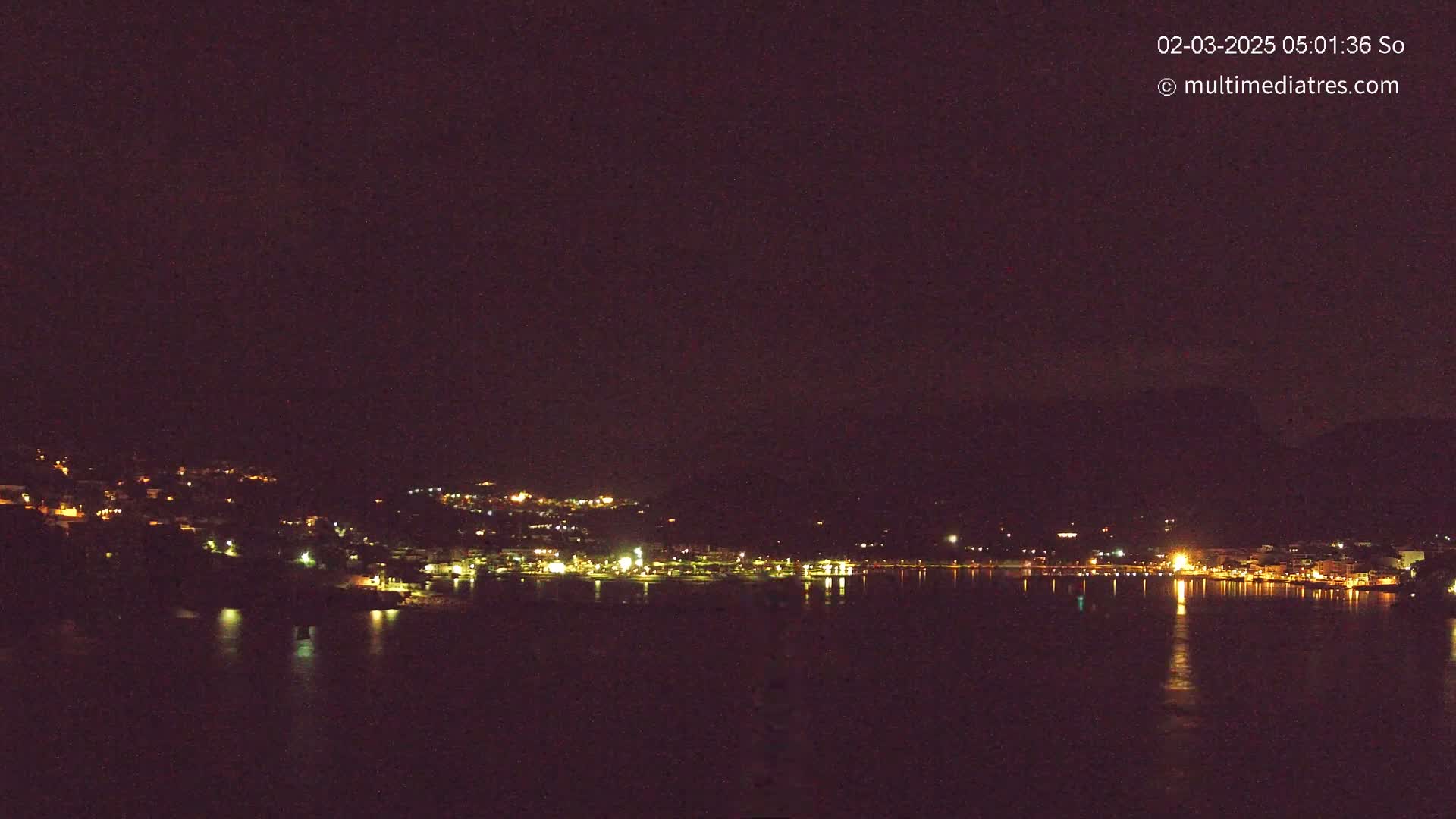 A nighttime view of a coastal town with its lights reflected in a calm body of water under a dark sky.