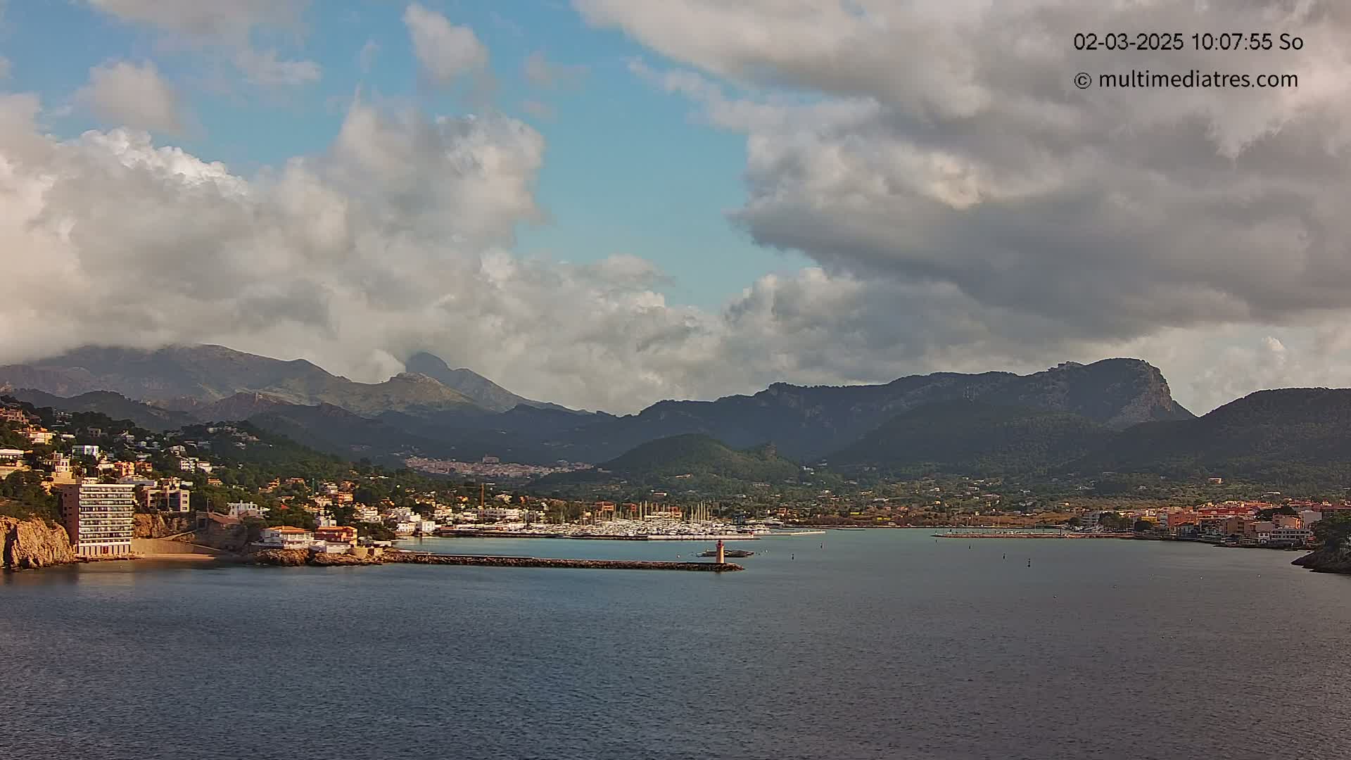 A coastal town nestled against a mountain range under a partly cloudy sky overlooks a calm harbor filled with boats.