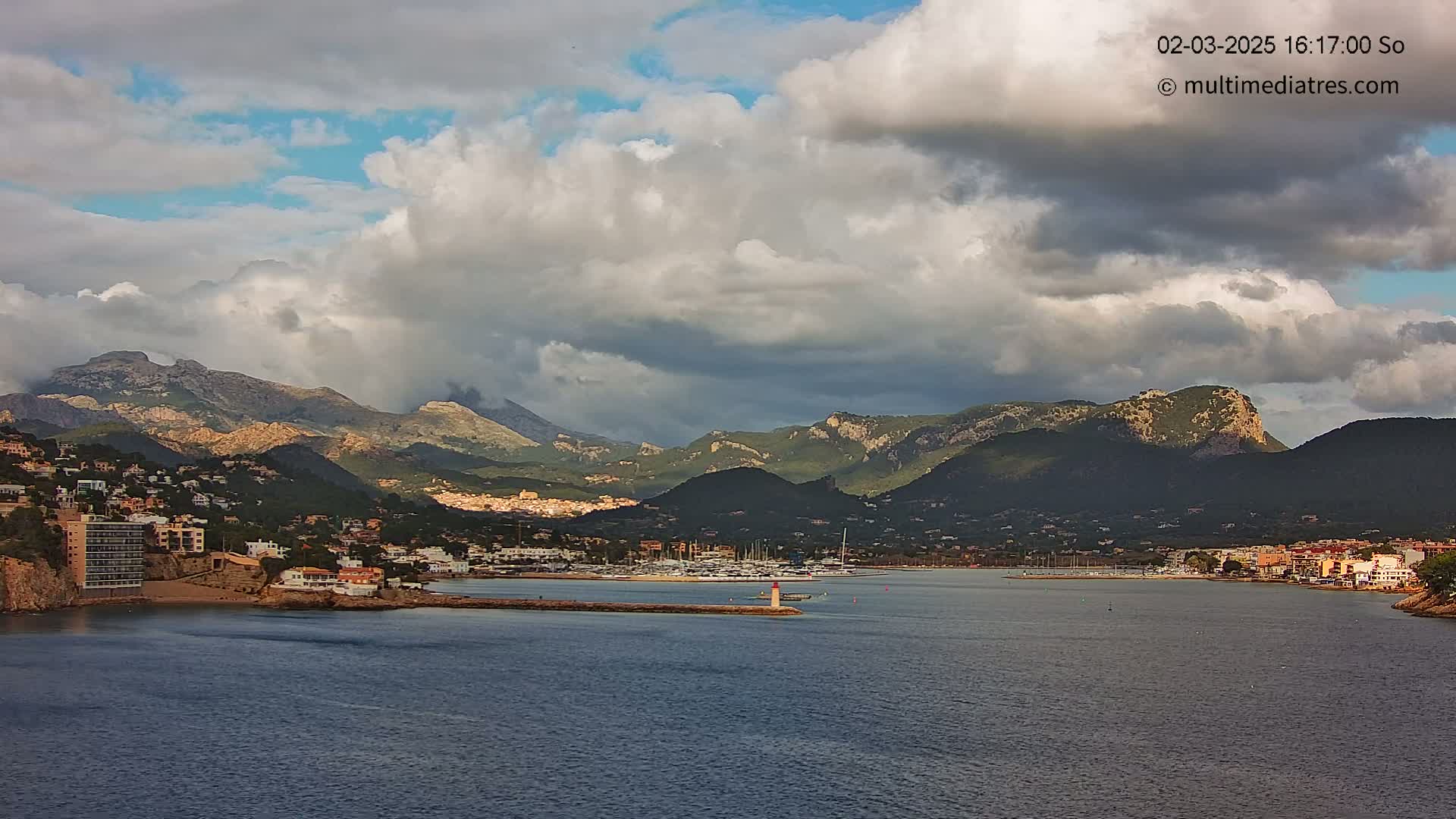 A coastal town nestled against a mountain range under a partly cloudy sky sits beside a calm body of water filled with boats.