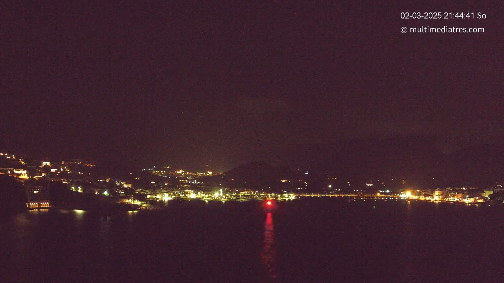 A nighttime view shows a coastal town illuminated with lights, situated on a hillside overlooking dark water; a single red light is visible on the water.