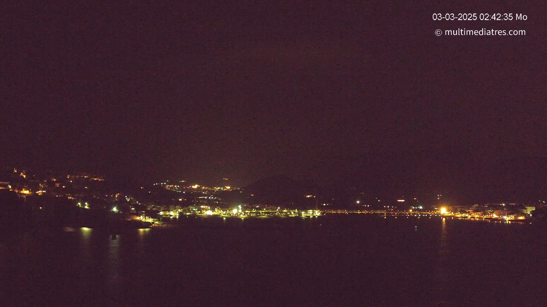 A nighttime view of a town's lights scattered along a dark hillside and reflected in a calm body of water under a clear, dark sky.