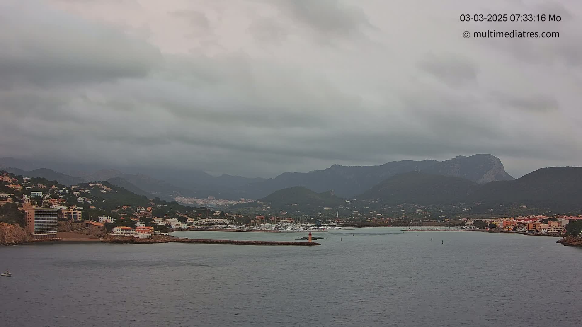 A coastal town nestled against a mountain range under a cloudy, overcast sky, with a calm body of water separating the town from the viewer's perspective.